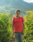 A man wearing a red polo shirt and blue jeans stands on a farm path surrounded by lush green plants, with mountains in the background under a cloudy sky.