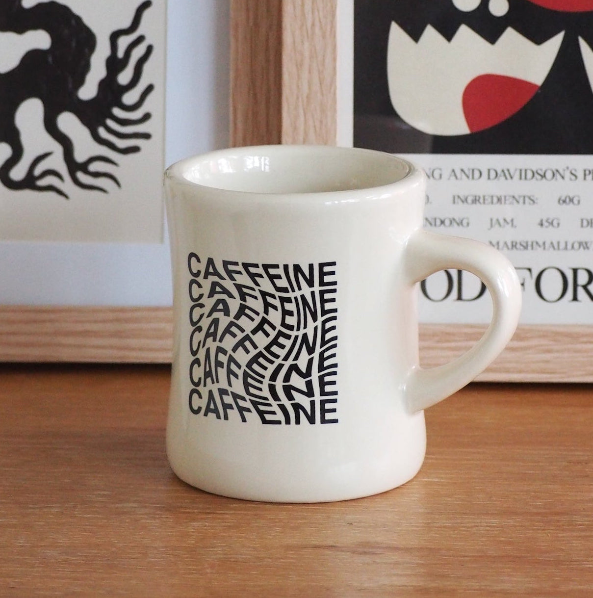 A white ceramic mug with black 'CAFFEINE' typography sits on a wooden table in front of framed wall art.