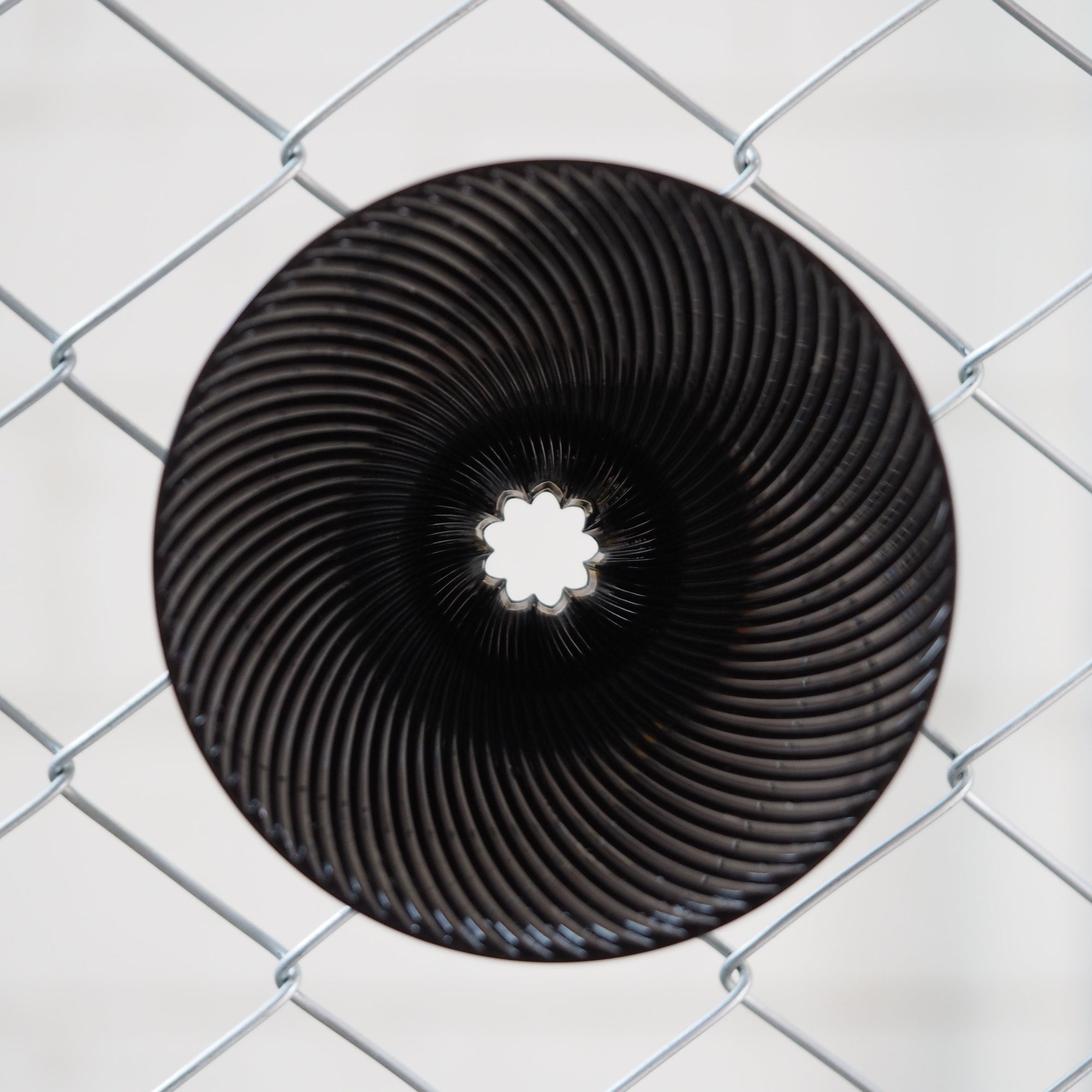 A circular black metal sculpture with concentric spiral ridges and a star-like opening in the center, photographed through a silver chain-link fence against a light gray background.