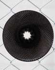 A circular black metal sculpture with concentric spiral ridges and a star-like opening in the center, photographed through a silver chain-link fence against a light gray background.